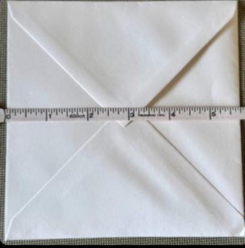 May include: A white envelope with a folded flap, measured with a tape measure. The envelope is approximately 5 inches wide. The envelope is on a neutral background.