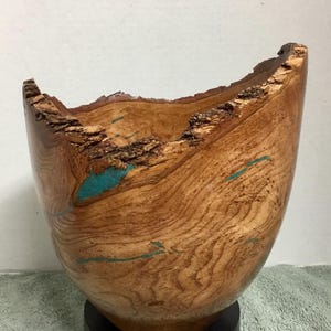 May include: A handcrafted wooden bowl with a natural edge and turquoise accents. The bowl is made of light brown wood with visible grain patterns and sits on a black base. The interior features turquoise-coloured inlays.