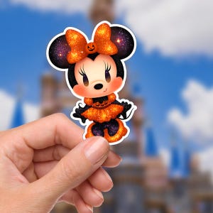 May include: A Halloween-themed sticker featuring a cartoon character with a pumpkin bow and dress. The character has black ears, a black body, and an orange and black outfit. The background is a blurred image of a castle and a blue sky.