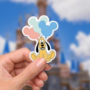 May include: A Pluto sticker holding three Mickey Mouse-shaped balloons in pastel colors. The sticker features a yellow Pluto with black ears and a blue collar. The background shows a blurred image of a castle and a blue sky.