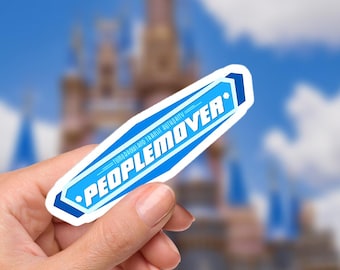 May include: A blue and white sticker with the text "PEOPLEMOVER" and "TOMORROWLAND TRANSIT AUTHORITY". The sticker is held in a hand, with a blurred background of a castle and blue sky.