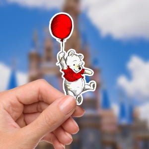 May include: A sticker featuring a cartoon bear holding a red balloon. The bear is wearing a red shirt and has a smiling expression. The background is a blurred image of a castle and a blue sky with clouds.