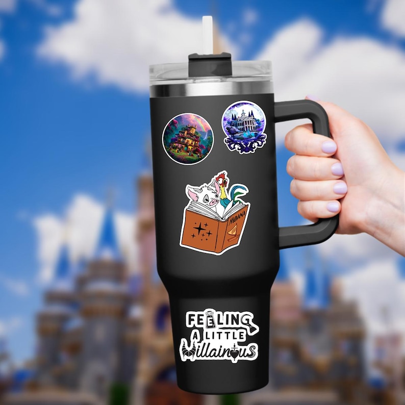 May include: A black insulated tumbler with a handle, adorned with stickers. The stickers feature a haunted house, a mansion, and characters from a storybook. The phrase "Feeling a little villainous" is also displayed.