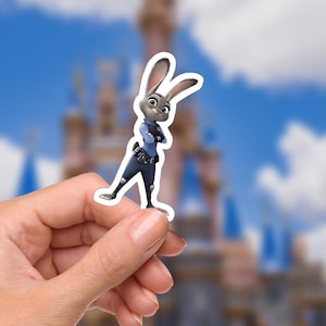 May include: A sticker featuring Judy Hopps from Zootopia. The sticker depicts Judy, a gray rabbit, in a police uniform with her arms crossed. The background is a blurred image of a castle and a blue sky with clouds.