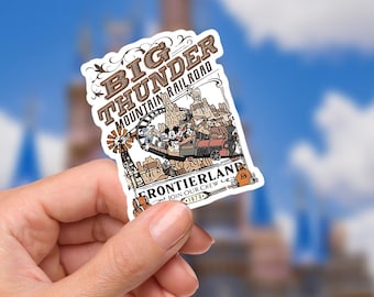 May include: A rectangular sticker featuring the Big Thunder Mountain Railroad ride at Frontierland. The sticker has a brown and white color scheme with Mickey Mouse and friends riding a train. The text "Big Thunder Mountain Railroad" and "Frontierland" are visible.