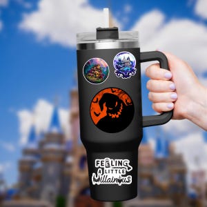 May include: A black tumbler with a handle, adorned with stickers featuring a haunted house, a mansion, and a silhouette of a character against an orange moon. The text "Feeling a little villainous" is also on the tumbler.