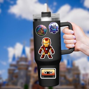 K&ouml;nnte beinhalten: Schwarzer isolierter Becher mit Griff, verziert mit Aufklebern von Iron Man, einem Spukhaus, einem Schloss und einer Retro-Kassette mit der Aufschrift "Awesome Mix Vol. 2". Der Becher hat einen transparenten Deckel und einen wei&szlig;en Strohhalm.