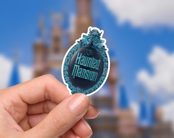 May include: A teal and black Haunted Mansion sticker. The oval-shaped sticker features the words "Haunted Mansion" in a stylized font, surrounded by a decorative teal border. The background is blurred, with a castle and blue sky.