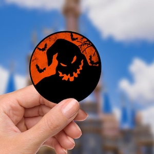 May include: A round black and orange sticker featuring a silhouette of a character with a sinister grin, bats, and a tree branch. The sticker is held in a person's hand against a blurred background of a blue sky and a castle.