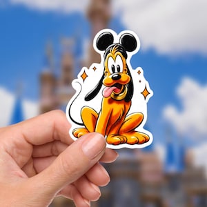 May include: A sticker featuring Pluto, the Disney cartoon dog, in a sitting pose with his tongue sticking out. The sticker has a white border and is held in a person's hand. The background is a blurred image of a castle.