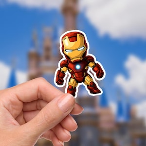 K&ouml;nnte beinhalten: Ein Aufkleber von Iron Man im Chibi-Stil. Der Aufkleber zeigt den Superhelden in seiner klassischen rot-goldenen R&uuml;stung mit einem blauen Lichtbogenreaktor. Der Hintergrund ist ein verschwommenes Bild eines Schlosses und eines blauen Himmels.