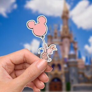 May include: A sticker featuring Piglet from Winnie the Pooh, holding onto a pink Mickey Mouse balloon. The cartoon character is light pink with a striped shirt. The background is a blurred image of a castle and blue sky.