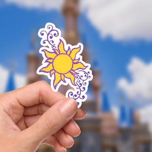 May include: A yellow sun sticker with purple accents and swirling vines is held in a hand. The sun has a smiling face and is surrounded by stylized rays. The background is a blurred image of a castle and a blue sky.