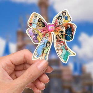 May include: A colorful bow-shaped sticker featuring various Disney princesses. The bow has a pink center and gold glitter edges. Each loop of the bow showcases a different princess, with a pink gem in the center. The background is a blurred image of a castle and blue sky.