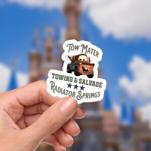 Pode incluir: Um autocolante com o Tow Mater do filme Carros. O autocolante tem o texto "Tow Mater" acima de uma imagem da personagem. Abaixo da imagem, o texto diz "Towing & Salvage, Radiator Springs" com três estrelas.