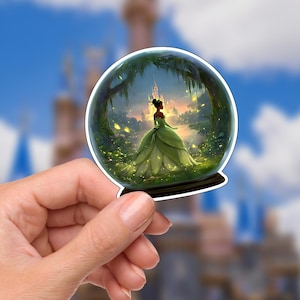 Pode incluir: Um autocolante com uma princesa num vestido verde dentro de um globo de neve. A princesa está numa floresta com um castelo ao fundo. O autocolante tem uma borda branca e uma base preta.