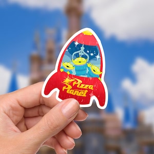 May include: A red rocket-shaped sticker with the Pizza Planet logo and three green aliens inside. The aliens are in a blue and white space scene. The sticker is held in a hand.