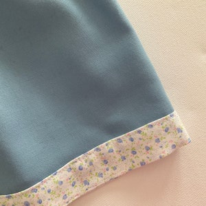 May include: A close-up of a light blue fabric with a floral patterned trim. The trim features small blue flowers and green leaves on a white background. The fabric appears smooth and soft.