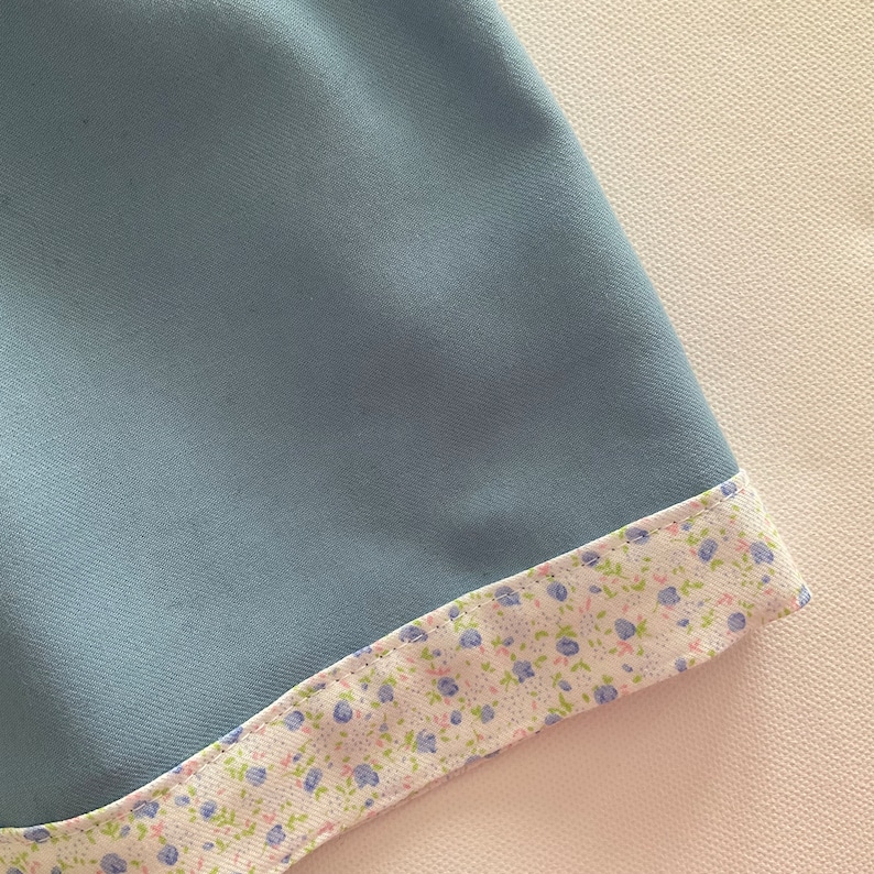 May include: A close-up of a light blue fabric with a floral patterned trim. The trim features small blue flowers and green leaves on a white background. The fabric appears smooth and soft.
