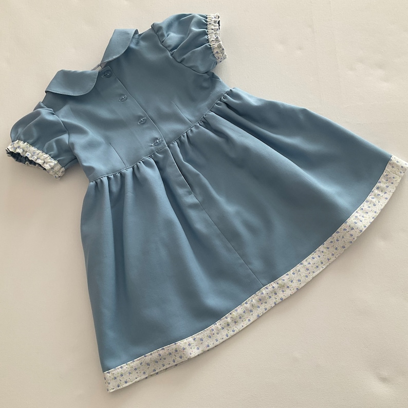 May include: A light blue baby dress with short, puffed sleeves and a Peter Pan collar. The dress features a button-down back and a gathered skirt. The hem and sleeve cuffs are trimmed with a white floral patterned fabric.