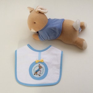 May include: A soft toy bunny in tan and blue, positioned above a white baby bib. The bib features a blue border, a circular rabbit illustration, and a yellow bow. The bib is designed for a baby.