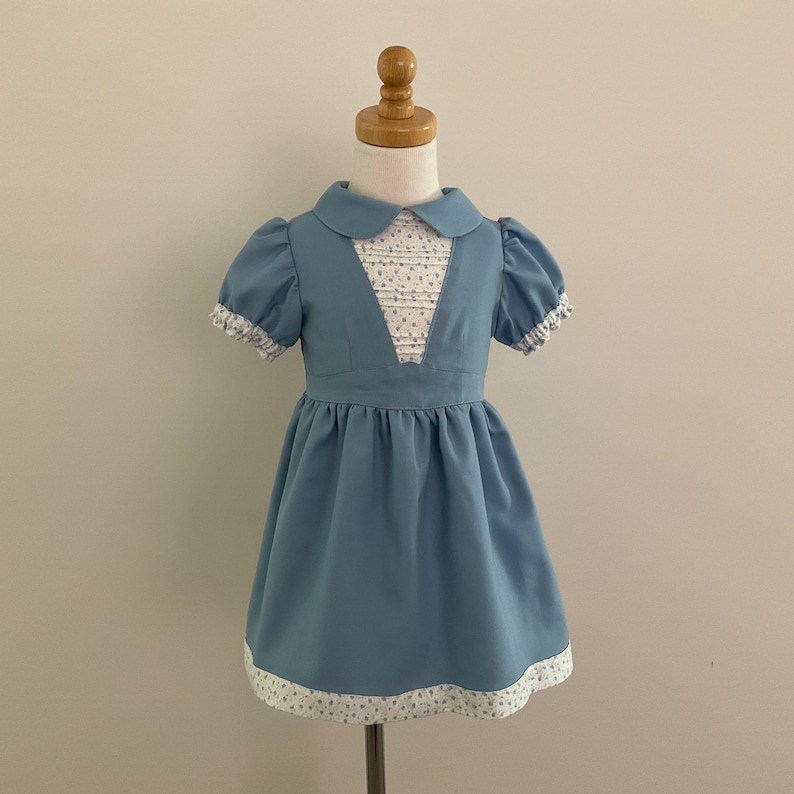 May include: A light blue dress for a child, displayed on a mannequin. The dress features a Peter Pan collar, short puffed sleeves, and a white bodice insert with a floral pattern. The hem and sleeve cuffs are trimmed with a matching floral fabric.