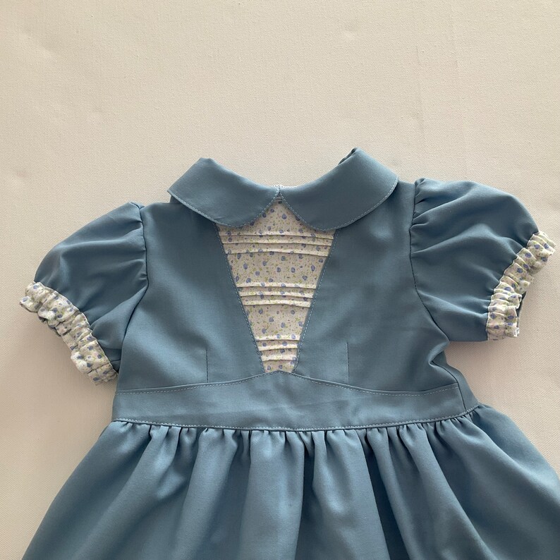 May include: A light blue dress with a Peter Pan collar and short, puffed sleeves. The bodice features a floral patterned panel with pleats. The sleeve cuffs and collar are trimmed with a floral print. The dress has a gathered skirt.