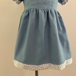 May include: A light blue dress with short sleeves and a gathered skirt. The hemline is trimmed with a white and floral patterned fabric. The dress is displayed on a metal stand.