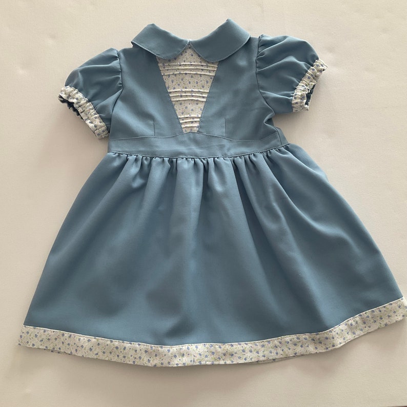 May include: A light blue baby dress with a Peter Pan collar and short, puffed sleeves. The bodice features a pleated insert with a floral pattern. The hem and sleeve cuffs are trimmed with a matching floral fabric. The dress has a gathered skirt.