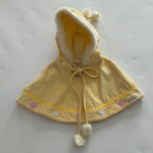 Pode incluir: Uma capa de veludo cotelê amarela com capuz forrado com pele sintética branca. A capa apresenta um fecho de cordão com pompons e é decorada com flores bordadas ao longo da bainha. O texto "mini bambini" é visível.
