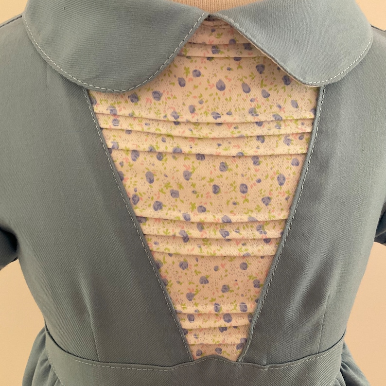 May include: A light blue dress with a Peter Pan collar and a pleated bodice insert. The insert features a floral pattern with blue and purple flowers on a cream background. The dress has short sleeves and a fitted waist.