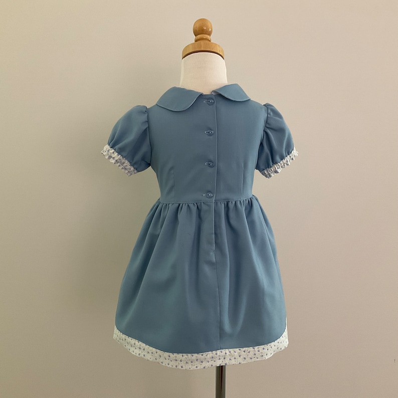 May include: A light blue dress with a Peter Pan collar and short, puffed sleeves. The dress features a button-down front and a gathered skirt. The sleeves and hem are trimmed with white lace detailed with a floral pattern. The dress is displayed on a mannequin.