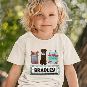 May include: A white t-shirt featuring a brown dog wearing a bow tie, surrounded by red crabs and blue coolers. The text "BRADLEY" is on a blue and white checked background.