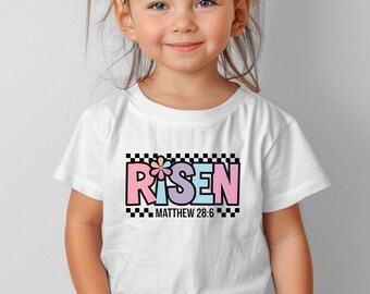 May include: White t-shirt with a colorful graphic that says "RISEN" with a flower in the "I". The graphic is surrounded by a black and white checkered border. The text "MATTHEW 28:6" is below the graphic.