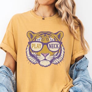 May include: Mustard yellow t-shirt featuring a tiger face graphic wearing sunglasses. The tiger graphic is purple and gold, with the words "PLAY NECK" printed on the design. The shirt has short sleeves and a relaxed fit.