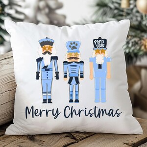 May include: White square pillow featuring three blue nutcracker figures with different hat designs and mustaches. The text "Merry Christmas" is written in a dark blue script below the figures. The pillow is on a wooden surface.