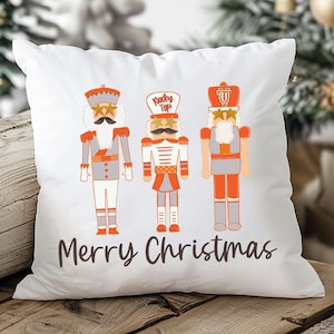 May include: White square pillow featuring three cartoon nutcracker soldiers in gray, orange, and white. The pillow has the text "Merry Christmas" in brown script. The pillow is on a wooden surface.