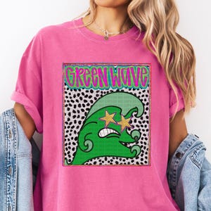 May include: A pink t-shirt featuring a graphic of a green wave with star-shaped eyes and the text "GREEN WAVE" in a neon green font. The wave design is set against a black and white polka dot background.