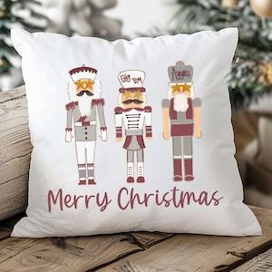 May include: White square pillow featuring three cartoon nutcracker figures in gray, red, and white. The words "Merry Christmas" are printed in a burgundy script below the figures. The pillow is on a wooden surface.