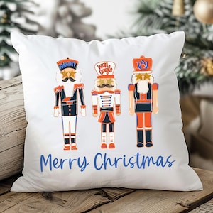 May include: White square pillow featuring three colorful nutcracker soldier illustrations. The nutcrackers are in red, blue, and orange uniforms. The words "Merry Christmas" are written in blue script across the bottom.