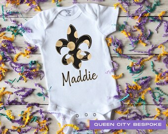 Puede incluir: Un body blanco para bebé con un diseño de flor de lis negro y dorado y el nombre "Maddie" impreso debajo. El body está rodeado de confeti morado, amarillo y verde.