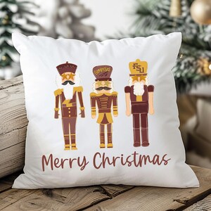 May include: White square pillow featuring three nutcracker figures in brown, gold, and maroon. The pillow has the words "Merry Christmas" in a maroon script across the bottom. The background includes a Christmas tree.