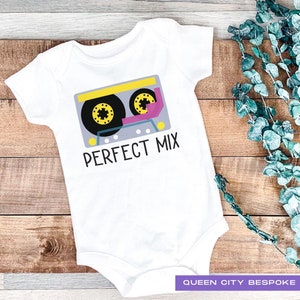 Puede incluir: Un body blanco para bebé con un gráfico de una cinta de casete y el texto "PERFECT MIX". El body tiene un diseño de cinta de casete amarillo, rosa y azul.