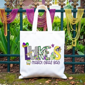 May include: White tote bag with the text "LUKE'S Mardi Gras Bag" in colorful letters with Mardi Gras themed decorations. The bag is hanging on a green metal fence with purple, gold, and green beads.