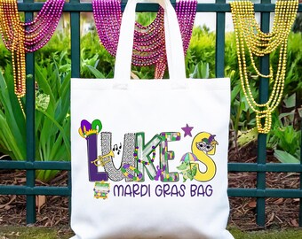 May include: White tote bag with the text "LUKE'S Mardi Gras Bag" in colorful letters with Mardi Gras themed decorations. The bag is hanging on a green metal fence with purple, gold, and green beads.