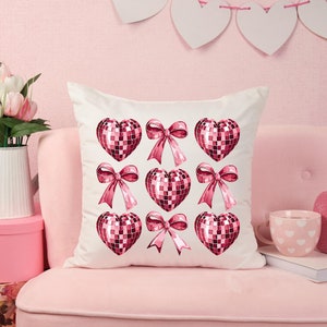 May include: White square throw pillow featuring a pattern of pink disco ball hearts and pink bows. The pillow is on a pink couch, with a pink background and decorative hearts hanging above.