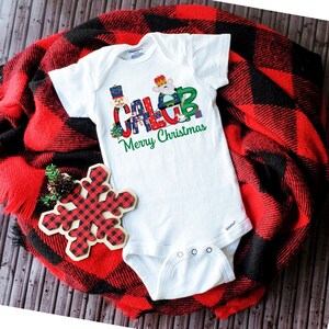 May include: White baby onesie with a Christmas-themed design featuring the name "Caleb" spelled out with festive characters and the text "Merry Christmas".