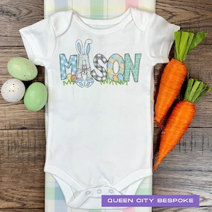 May include: White baby onesie featuring a blue and green Easter bunny and the name "MASON" in a colourful patchwork design.