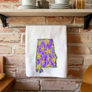 Könnte beinhalten: Ein weißes Küchentuch mit einer lila und goldenen Umrandung des Bundesstaates Alabama mit goldenen Sternen.