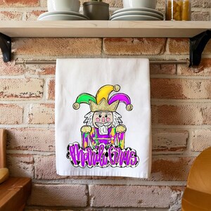 Może przedstawiać: Biały ręcznik kuchenny z ilustracją karnawałowego błazna w kolorze fioletowym, zielonym i złotym oraz napisem "Mardi Gras".